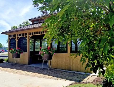 Wooden gazebo beside a paved path, leafy trees and hanging flower pots