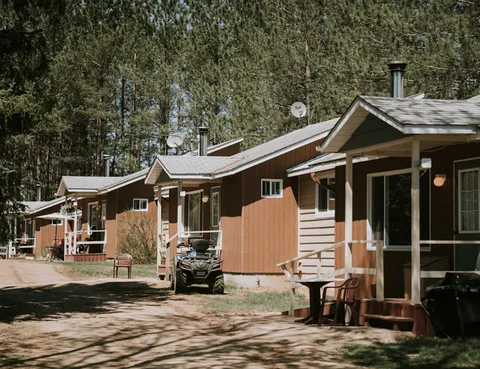 Row of single-storey brown cottages with porches beside a wooded lane