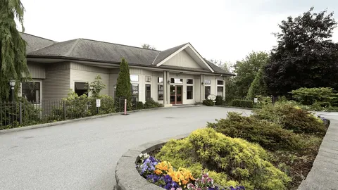 Residence exterior with an entrance, a paved driveway, flower beds, shrubs and trees.