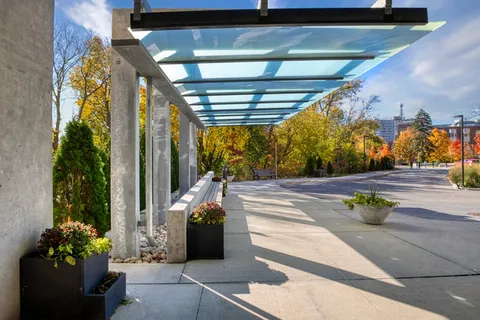 Glass canopy over a paved walkway with flower planters beside a driveway