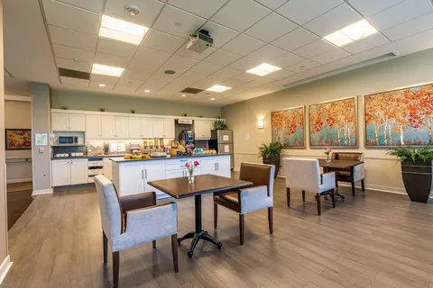 Tables and upholstered chairs beside a white kitchen and tree paintings