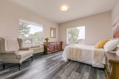 Bedroom with a bed, patterned armchair, wooden dresser and two windows