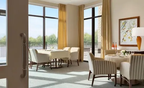 Dining area with tables and upholstered chairs beside tall windows.