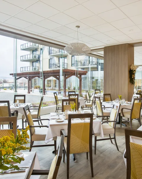 Dining tables with white linens and yellow-backed chairs beside tall windows