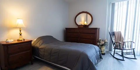 Bedroom with a grey bedspread, wooden dresser, round mirror and rocking chair