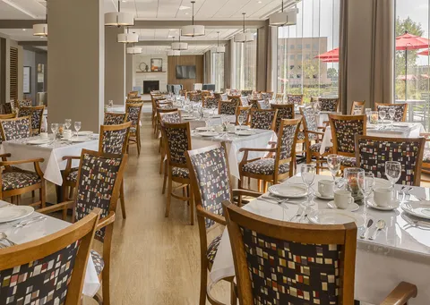 Dining tables set with white linens, glassware and patterned chairs beside tall windows