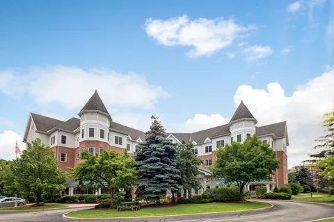 Residence with turreted corners beside lawns, trees and an entrance drive.