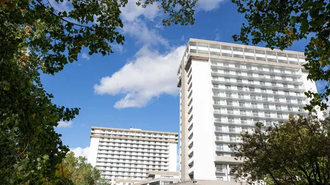White residence tower with rows of balconies, viewed through trees.