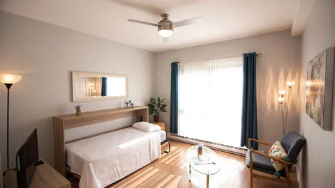 Studio with an open wall bed, chairs and a balcony door.