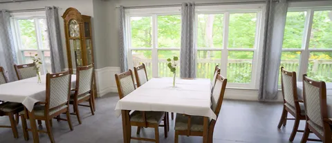 Tables with white cloths and wooden chairs beside large windows and a tall wooden clock