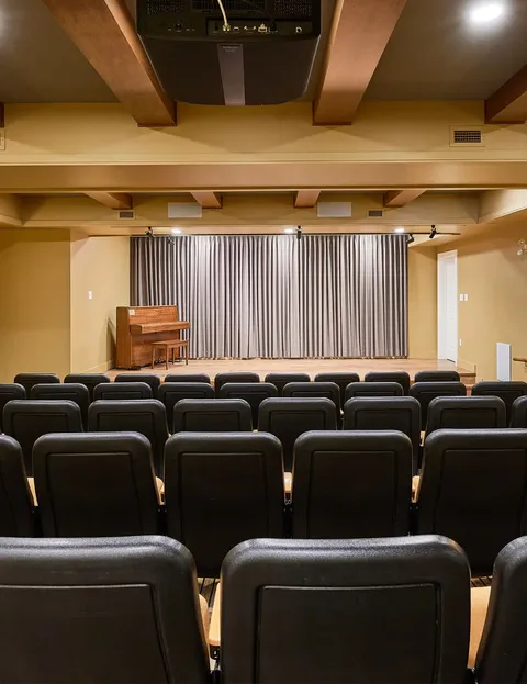 Theatre with rows of seats, a piano and closed stage curtains.