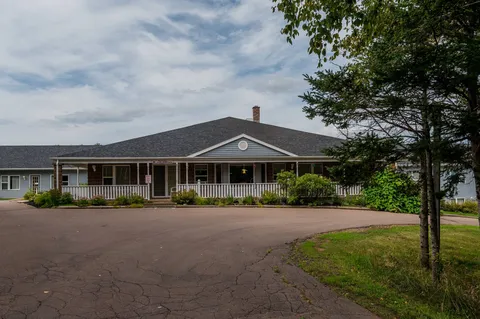 Long low-rise building with a covered porch beside a paved drive and mature tree