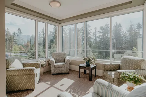 Sunroom with upholstered chairs, a small table, plants and windows overlooking trees.