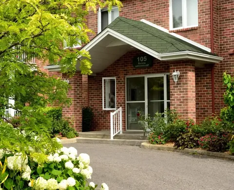 Brick entrance with glass doors, a covered porch and surrounding flowers and trees