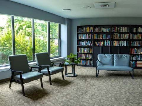 Library seating with blue chairs, a loveseat, bookshelves and large windows