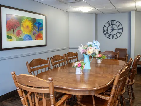 Long wooden dining table and chairs beneath a flower painting and wall clock