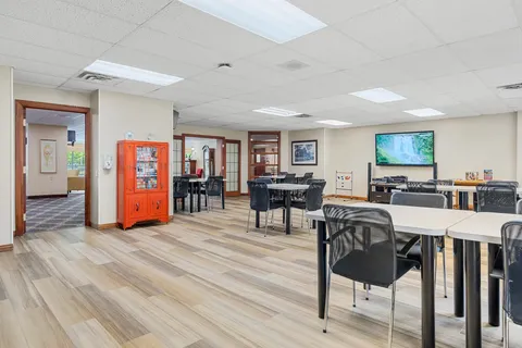 Room with tables, mesh-backed chairs, bookshelves and a television showing a landscape