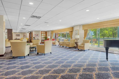 Seating area with cream armchairs, tables, a piano and large windows