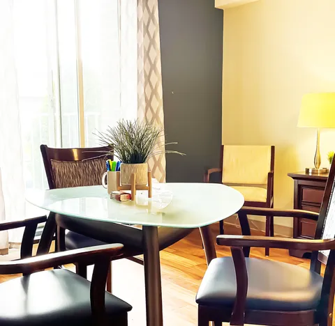Sitting area with a round glass table, wooden chairs and a potted plant.