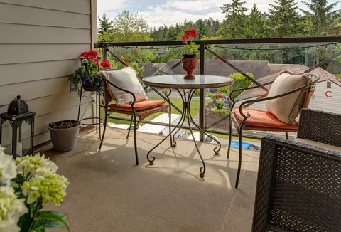 Covered balcony with a glass-top table, cushioned chairs, flower pots and a railing.