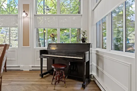 Upright piano and stool beside tall windows and potted plants