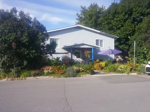 White-sided residence behind a flower garden, with blue porch posts and a purple patio umbrella