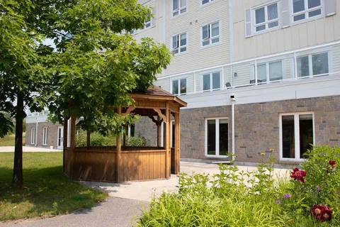 Side of a residence beside a wooden gazebo, lawn, paved path and flower beds