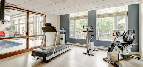 Exercise room with a treadmill and bicycles beside windows overlooking a pool