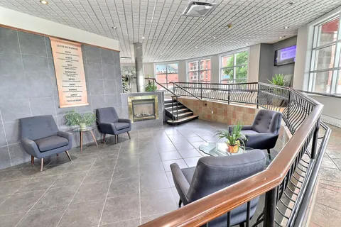 Lobby with armchairs, plants, a fireplace and ramp beside tall windows