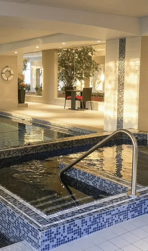 Indoor pool and raised hot tub with mosaic tiles, a metal handrail and potted trees