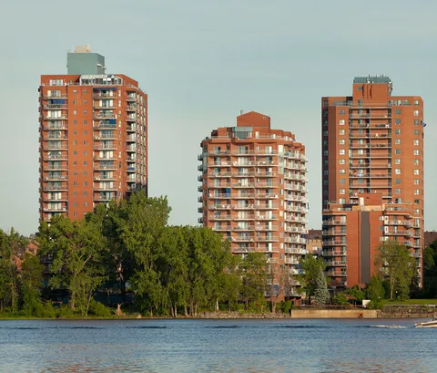 Three brick residential towers with balconies beside trees and the river