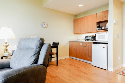 Kitchenette with wood cupboards, a mini-fridge and microwave beside a grey recliner