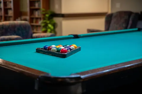 Green pool table with racked billiard balls, armchairs and bookshelves behind it
