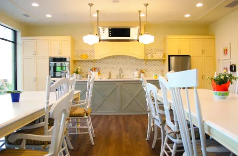 Kitchen with yellow cabinets, an island, wall ovens and white tables and chairs