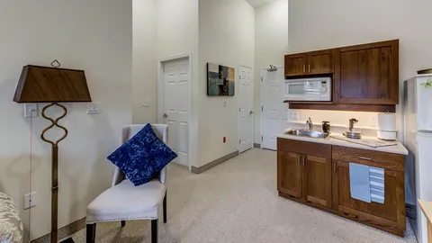 Room with an upholstered chair, a lamp and a kitchen area with wooden cupboards