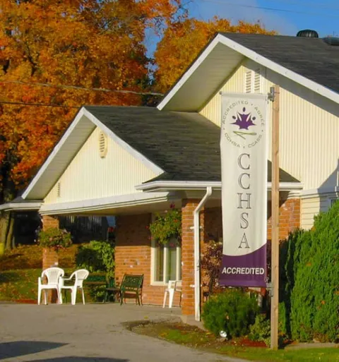 Covered entrance beside a brick wall, shrubs and outdoor chairs.