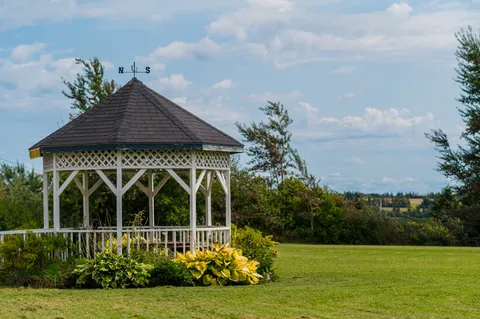 White gazebo with a shingled roof beside a lawn and flowering plants