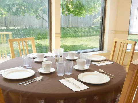 Dining table set with plates, cups and glasses beside garden-facing windows.