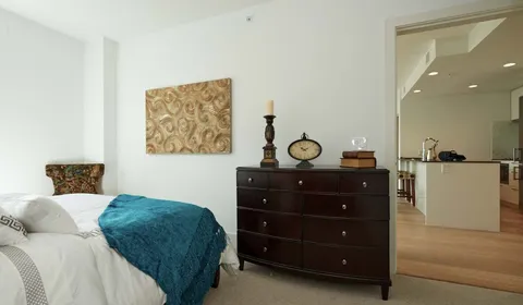 Bedroom corner with wooden drawers and a clock beside a kitchenette