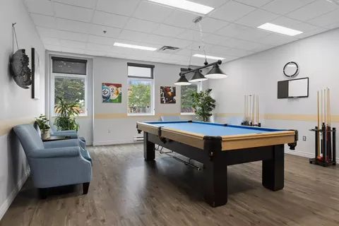 Billiards table beneath hanging lights beside armchairs and windows