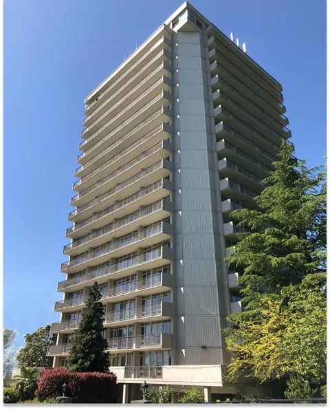 Tall residence tower with rows of balconies beside trees and planted beds