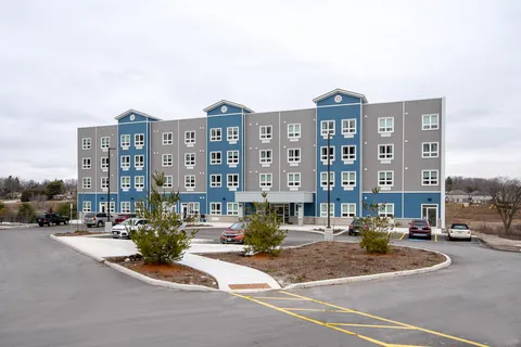 Grey and blue residence building with a curved drive, parked cars and small trees