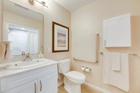Bathroom with a white vanity, toilet, grab bars and a mirror reflecting a shower