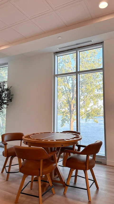 Round game table and wooden chairs beside tall river-facing windows.