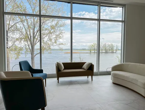 Sitting area with blue and cream chairs beside floor-to-ceiling river-facing windows.