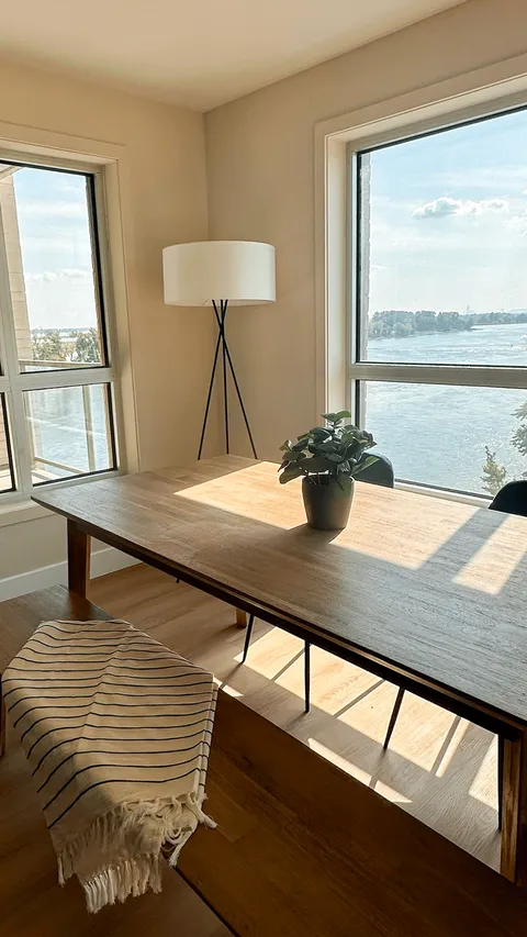 Wooden table, chairs and a floor lamp beside large river-facing windows.